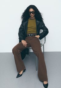 Bruine uitlopende broek, groene aansluitende top, zwarte leren jas en zwarte puntige hakken; model zittend op een metalen stoel voor een lichte achtergrond.