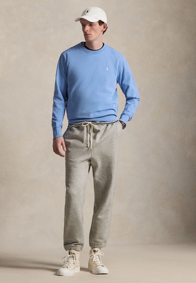 Άνδρας που φορεί ανοιχτό μπλε φούτερ, γκρι φόρμα, λευκά ψηλά αθλητικά παπούτσια και λευκό καπέλο μπέιζμπολ, στέκεται μπροστά σε ουδέτερο φόντο.