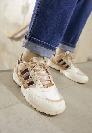 Personne portant des baskets Adidas beiges et blanches avec des bandes marron et un jean bleu retroussé, debout sur des escaliers en béton.