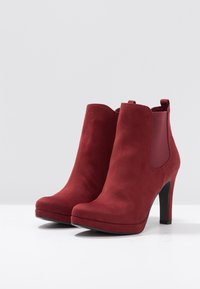Red suede ankle boots with a high heel, elastic side panels, and a smooth texture. Rounded toe and minimal design details.