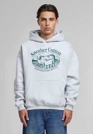 Hirose en coton gris avec une poche avant et un logo imprimé vert d'une vache avec le texte « Another Cotton » et « COTTON MANUFACTURER - SINCE 2023 ».