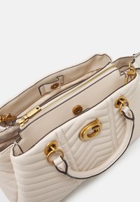 Sac à main en cuir matelassé beige avec quincaillerie en ton doré, fermeture à glissière, bouton-pression et poignées supérieures sur un fond blanc.