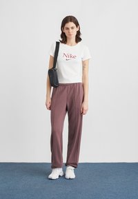 Λευκό cropped t-shirt με κόκκινο λογότυπο "Nike", συνδυασμένο με φαρδιά μπορντό παντελόνια και λευκά αθλητικά παπούτσια. Το μοντέλο κρατά μια μικρή μαύρη τσάντα.
