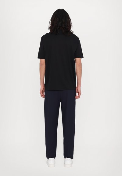 Tricou scurt cu mânecă neagră, cu o croială lejeră, purtat cu pantaloni bleumarin și adidași albi, văzut din spate. Textură netedă, fără modele.