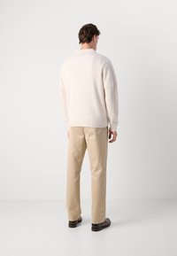 Crème kleurige trui met ronde hals en textuur, gecombineerd met lichtbeige broeken. Model staat met de rug naar de kijker tegen een lichte achtergrond.