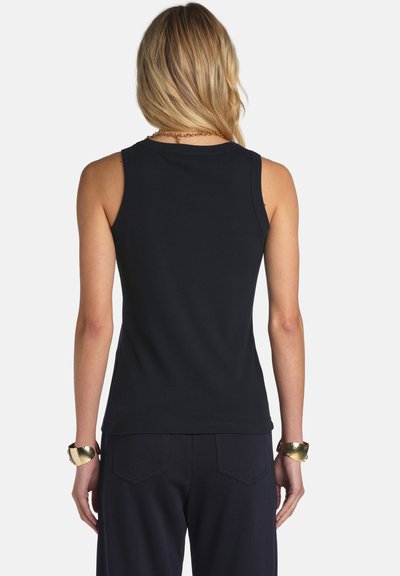 Szőke nő fekete ujjatlan felsőt és sötét nadrágot visel, hátulról nézve, arany karkötőkkel és nyaklánccal díszítve.