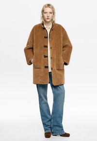 Abrigo de piel sintética marrón con un corte relajado, frente de botones y grandes bolsillos de parche, combinado con jeans azul de pierna recta.