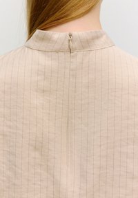 Hellbeige Kleidungsstück mit hohem Kragen, das zarte vertikale Nadelstreifen aufweist, einen Reißverschluss hinten hat und ein kleines Schlitzdetail am Saum besitzt.
