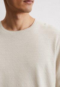 Beige gebreide trui met een ronde hals. De stof heeft een subtiel gebreid patroon, wat zorgt voor een zachte maar gestructureerde uitstraling.