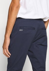 Homme portant un pantalon bleu marine avec des poches passepoilées à l'arrière et un petit logo blanc de ciseaux près de la poche droite, associé à une chemise blanche.