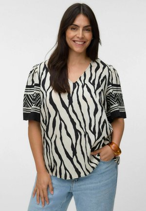 Femme souriante aux longs cheveux foncés portant un chemisier à imprimé zèbre noir et blanc et un jean bleu clair, une main dans la poche.
