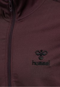 Chaqueta de punto burdeos con cremallera, hecha de un tejido suave, que presenta un logo bordado negro en el pecho y un detalle de costura minimalista.