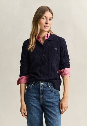 Donna che indossa un maglione lavorato a trecce blu navy sopra una camicia rosa con le maniche arrotolate e jeans blu, in piedi davanti a uno sfondo neutro.