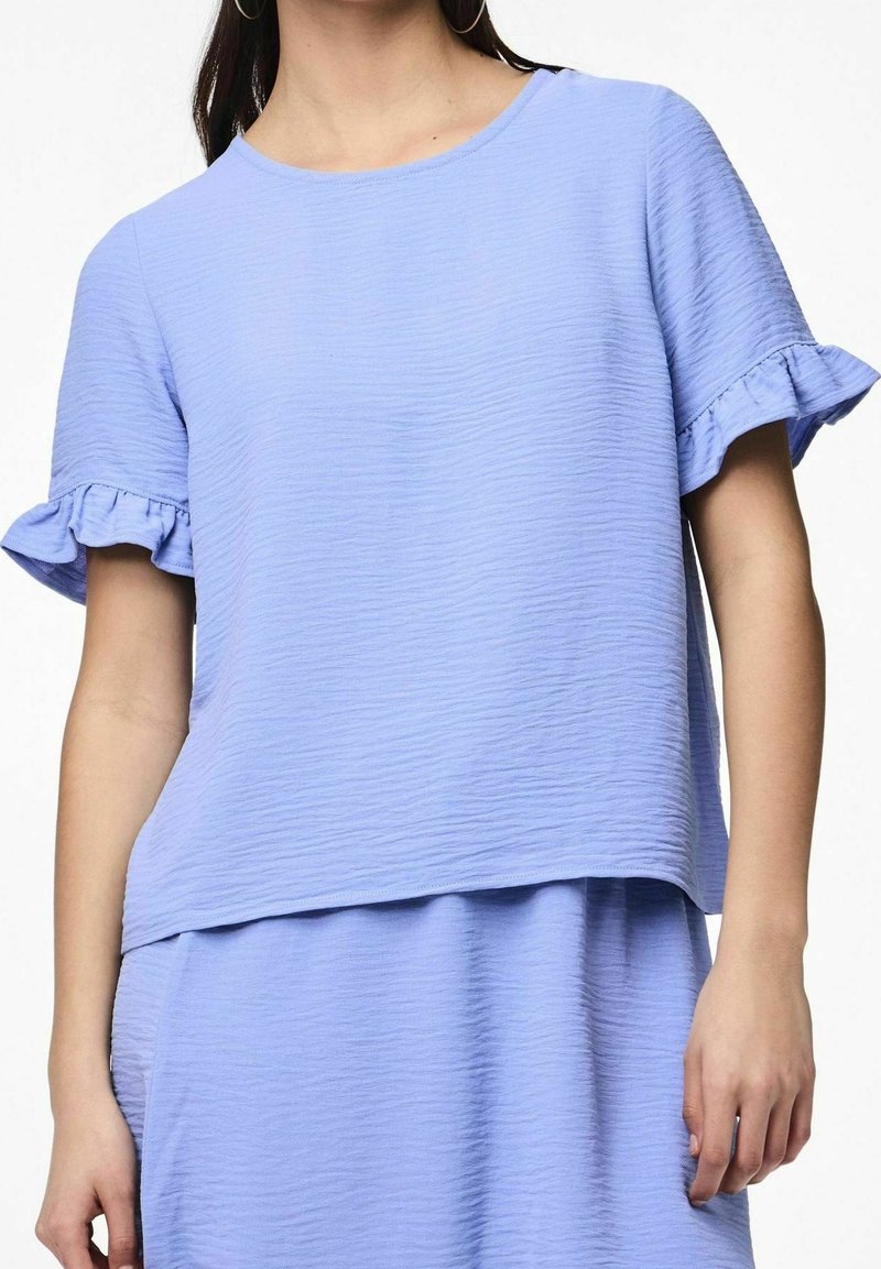 Femme portant un haut texturé bleu clair à manches courtes avec bords de manches volantés et une jupe assortie, visible du cou aux hanches.