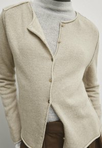 Cardigan in maglia beige con scollatura tonda, caratterizzato da bottoni lungo la parte anteriore. Presenta un tessuto strutturato e dettagli a costine.