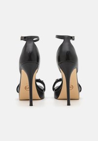 Černé sandály na vysokém podpatku s lesklým koženým povrchem, řemínkem kolem kotníku a tenkým stiletto podpatkem. Mají béžový interiér a detail loga.