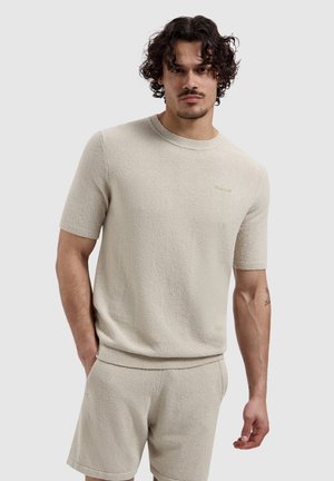 Jonge man met krullend haar die een beige gestructureerd T-shirt met korte mouwen en bijpassende korte broek draagt, staand met één hand in zijn zak.