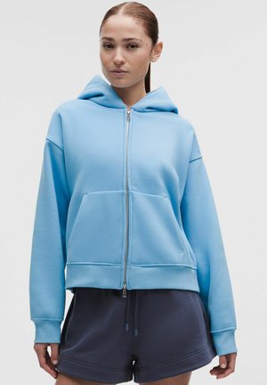 Kobieta z brązowymi włosami związanymi do tyłu, ubrana w jasnoniebieską bluzę z kapturem na zamek i ciemnoszare spodenki, stoi na jednolitym tle.