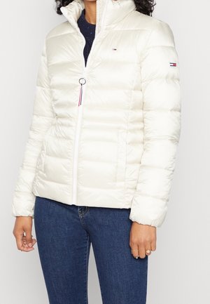 Femme portant une doudoune matelassée blanche avec un col montant et un jean bleu, montrant les logos devant et sur la manche.