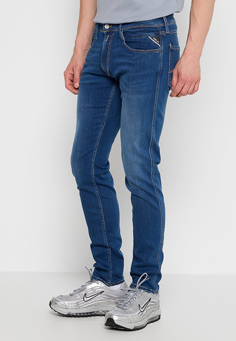 Męskie niebieskie dżinsy o slim fit, z pięcioma kieszeniami, widocznym stębnowaniem i delikatnym przetarciem. Połączone z srebrnymi butami sportowymi.