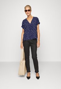 Blusa azul-marinho de manga curta com decote em V e padrões geométricos brancos, combinada com calças justas pretas, sapatos pretos de bico fino e uma mala bege.