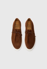 Une paire de chaussures en suède marron à enfiler avec des lacets en cuir et des semelles en caoutchouc blanc, vue de dessus sur un fond uni.