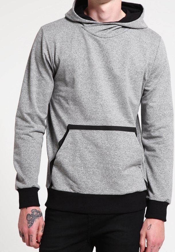 Man draagt een grijze hoodie met zwarte bies aan de zak, zwarte manchetten en zwarte broek, waarbij getatoeëerde handen zichtbaar zijn.