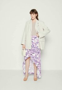 Woman wearing oversized white blazer, beige top, purple and white patterned asymmetrical skirt, tan heels, and holding a small white handbag.