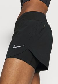 Svarta träningsshorts med inbyggd mesh-foder, utrustade med en dragkedjeficka och en silverfärgad Nike-logotyp på sidan. Designade för komfort och rörlighet.