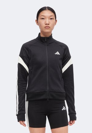 Joven con chaqueta negra con cremallera y pantalones cortos a juego con rayas blancas y logotipos de Adidas, de pie frente a un fondo liso.