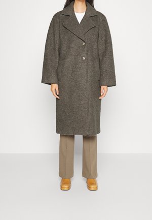 Femme portant un long manteau en laine gris foncé avec de larges revers, un pantalon beige et des chaussures en cuir marron clair, debout devant un fond blanc uni.
