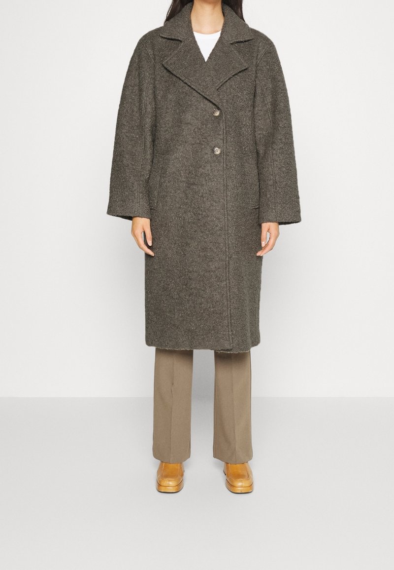 Femme portant un long manteau en laine gris foncé avec de larges revers, un pantalon beige et des chaussures en cuir marron clair, debout devant un fond blanc uni.