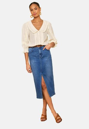 Frau trägt eine cremefarbene, strukturierte Bluse mit großem Kragen und Puffärmeln, einen blauen Denim-Midirock mit Frontschlitz, einen braunen Gürtel und braune Sandalen.