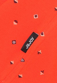 Rood gebreid stof met kleine geometrische patronen in marineblauw en wit. Zwarte label met "J&JOY" in witte stiksel op de rand.