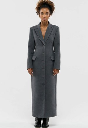 Kobieta o kręconych włosach nosi długi, dopasowany szary płaszcz na guziki z klapami i kieszeniami z patkami, stoi na tle jednolitego, jasnego tła.