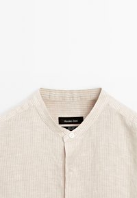 Leinenhemd in hellem Beige mit einer strukturierten Musterung, ausgestattet mit einem runden Kragen und einem einzelnen Knopfverschluss. Markenetikett innen sichtbar.