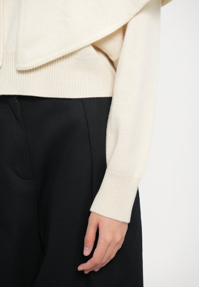 Un pull en tricot couleur crème avec un large col et des poignets côtelés, associé à un pantalon sombre. Le tissu semble doux et légèrement texturé.