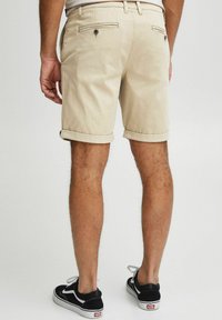 Shorts de algodón beige con puños enrollados, con bolsillos traseros y detalles en metal oscuro, combinados con zapatillas negras y blancas.