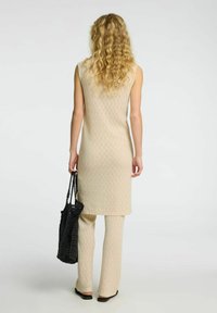 Robe sans manches tricotée beige avec un motif en chevrons, associée à un pantalon assorti. Sac fourre-tout en crochet noir. Texture lisse, silhouette ajustée.