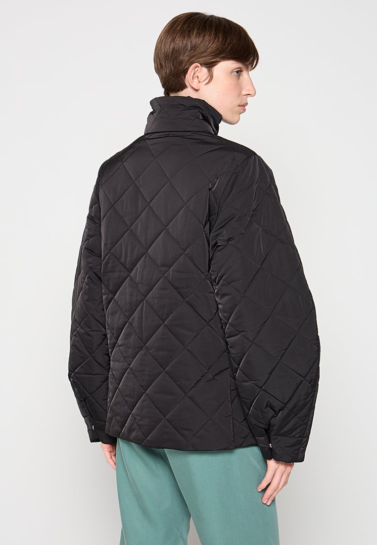 Zwarte gewatteerde jas met een hoge kraag, oversized silhouet en contrasterende stiksels. Zachte textuur met een matte afwerking.