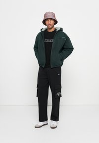Green fleece-lined jacket, black graphic t-shirt, black cargo pants with pockets, and a pink bucket hat. White sneakers with brown soles.