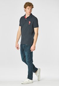Polo shirt navy con colletto corallo a contrasto, con logo sul lato sinistro del petto; abbinato a jeans blu scuro e sneakers bianche.