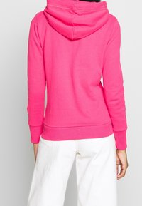 Rosa Kapuzenpullover mit weicher Textur, ausgestattet mit einer großen Kapuze, gerippten Bündchen und einem Saum, getragen mit weißen weiten Hosen.