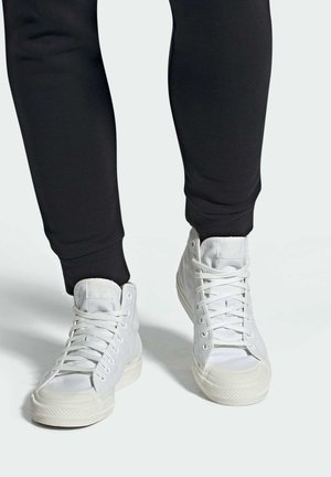 Zapatillas altas blancas con parte superior de lona, suela de goma y cordones clásicos, combinadas con joggers negros ajustados.