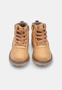 Stivaletti in pelle tan con lacci marroni, suola in gomma e zip laterale. Presentano una punta arrotondata e un colletto imbottito.
