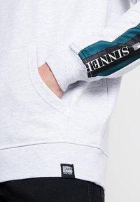 Personne avec la main dans la poche d'un sweat gris clair, comportant une rayure bleu-vert et noire avec le texte "TENNIS" sur la manche et une étiquette "SUPPLY & DEMAND" près de l'ourlet.