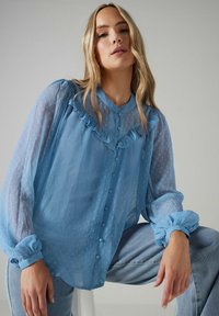 Blusa de gasa azul claro con un escote con volantes, frente abotonado y patrón de puntos texturizados. Presenta mangas abullonadas con puños de elastano.