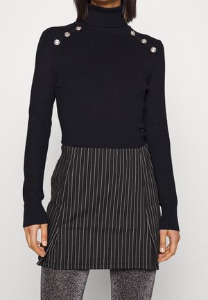 Femme portant un pull à col roulé noir avec des boutons argentés aux épaules et une jupe noire à fines rayures.