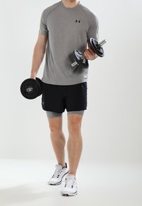 Muž v šedé košili a černých šortkách zvedající činky, stojící na světlém pozadí, oblečený v bílých sportovních botách.