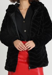 Černá bunda s kožešinovým podšívkou na zip, s chevronovým vzorem. Nosená přes černou košili, v kombinaci s červenou koženou sukní.